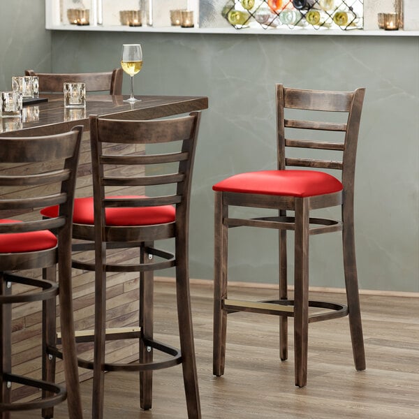 A Lancaster Table & Seating wood ladder back bar stool with a red vinyl seat next to a table with a glass of wine.