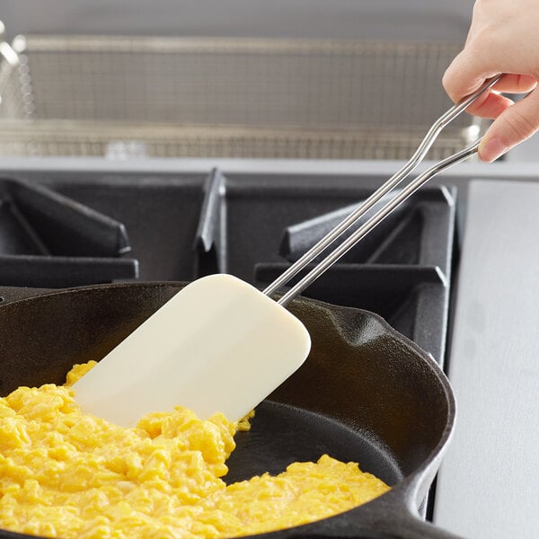A white Linden Sweden silicone spatula with a stainless steel handle cooking scrambled eggs in a pan.
