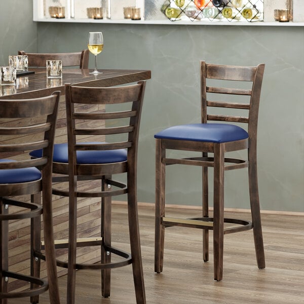A Lancaster Table & Seating wood ladder back bar stool with a detached navy vinyl seat on a table next to a glass of wine.