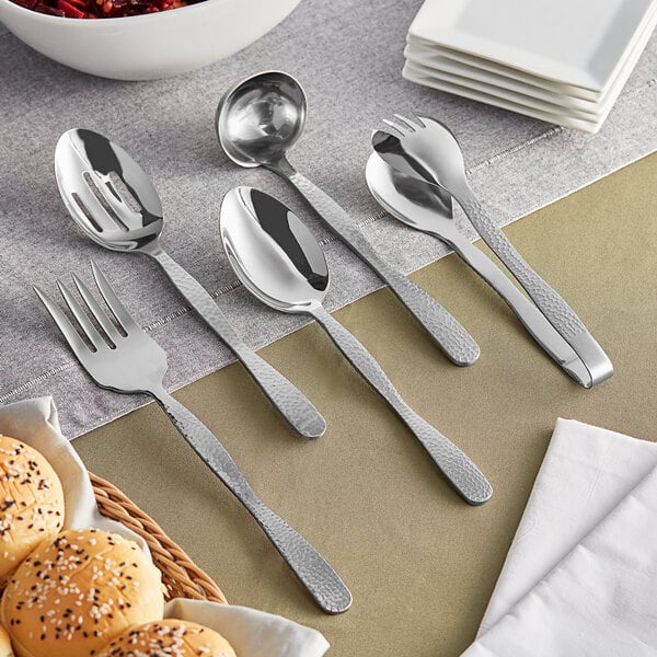 A table set with American Metalcraft stainless steel serving utensils, including a spoon and fork on a napkin with a basket of bread.