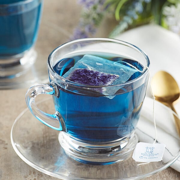 A glass cup with Wild Hibiscus Blue-Tee Butterfly Pea Flower Herbal Tea and ice cubes on a saucer.