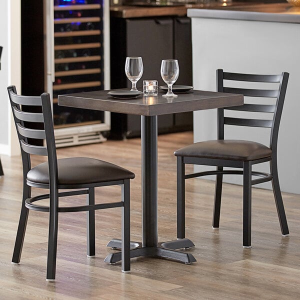 A Lancaster Table & Seating wood table with two chairs, wine glasses, and a candle on it.