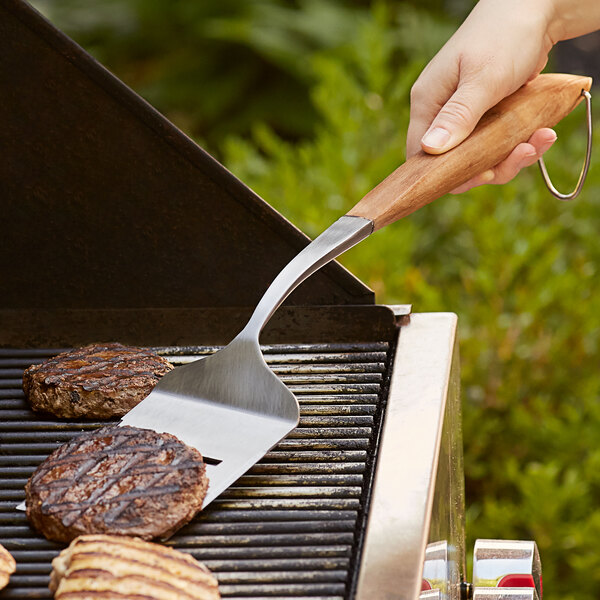 A person using an Outset stainless steel turner to cook meat on a grill.
