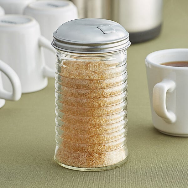 A glass beehive sugar pourer with brown sugar in it next to a white coffee mug.