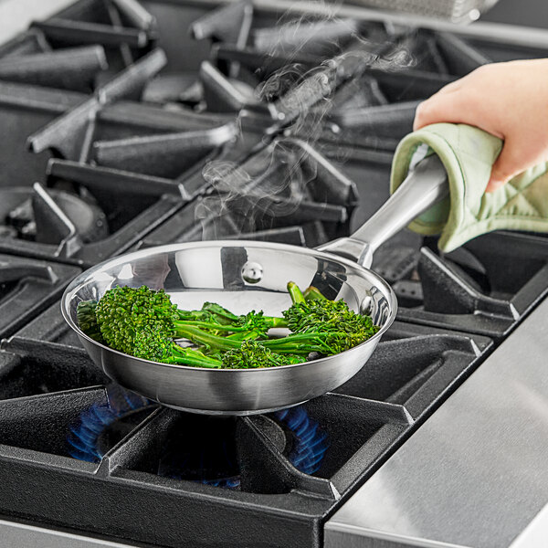 A hand using a spatula to cook broccoli in a Vollrath stainless steel fry pan.