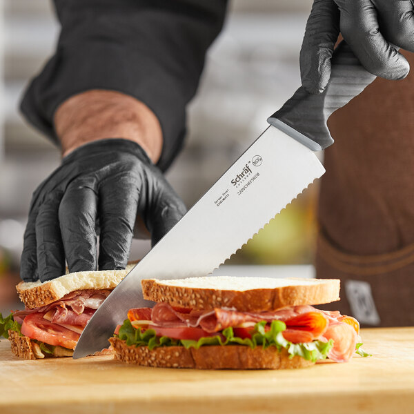 A person in black gloves using a Schraf serrated chef knife to cut a sandwich with meat and lettuce.