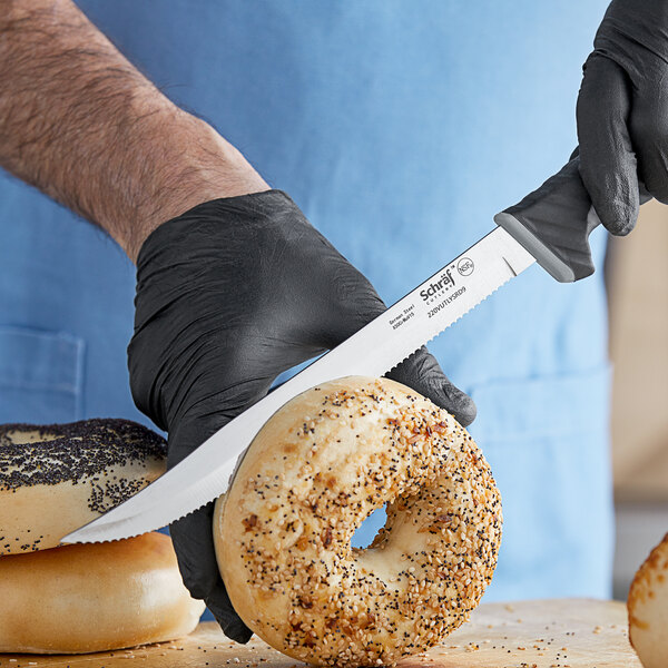A person in black gloves uses a Schraf serrated utility knife to cut a bagel on a counter.