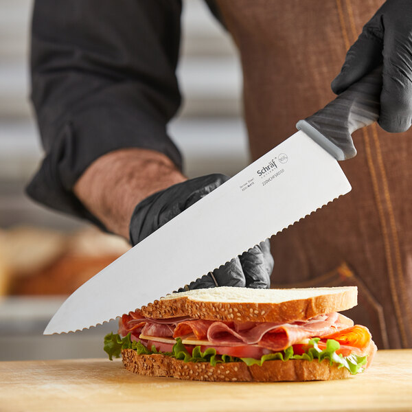 A person in black gloves uses a Schraf serrated chef knife to cut a sandwich with meat and lettuce.