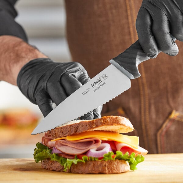 A person wearing black gloves uses a Schraf serrated chef knife to cut a sandwich.