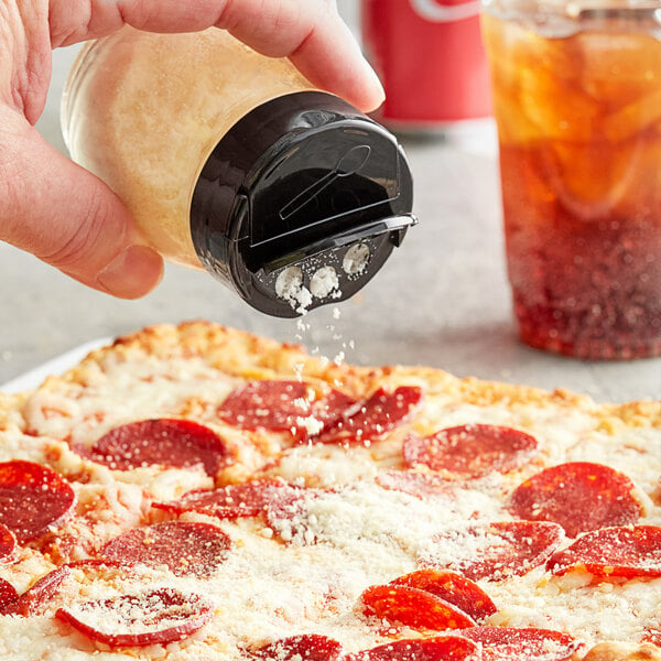 A pepperoni pizza being sprinkled with cheese from a Tablecraft clear glass shaker with a black plastic flip top.