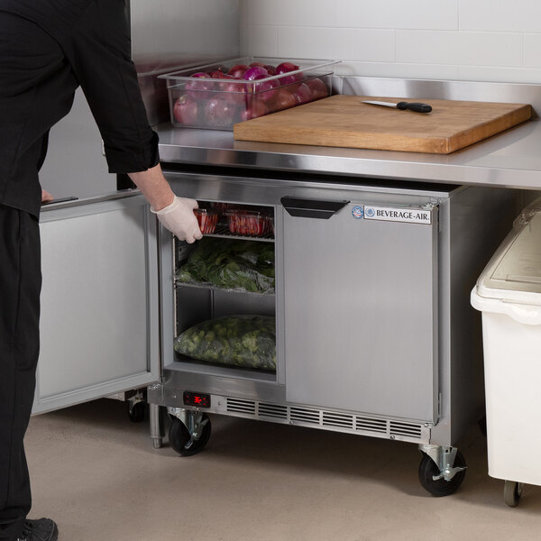 A woman using a Beverage-Air undercounter refrigerator on a counter in a school kitchen to put vegetables in a white container.