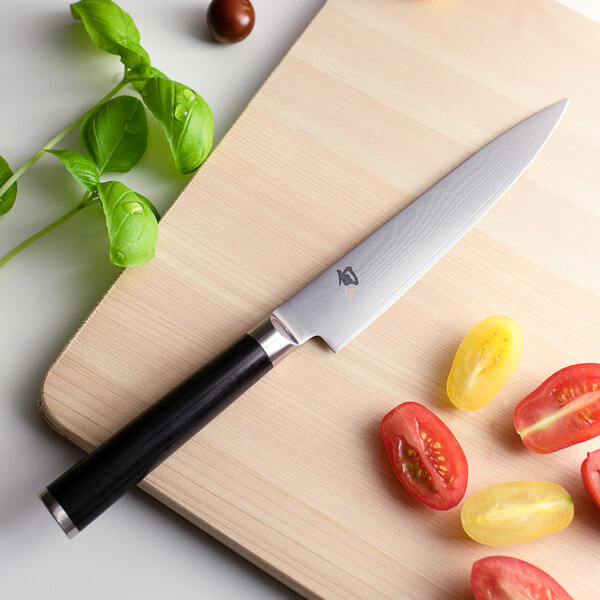 A Shun Classic forged utility knife cutting a yellow tomato on a cutting board.