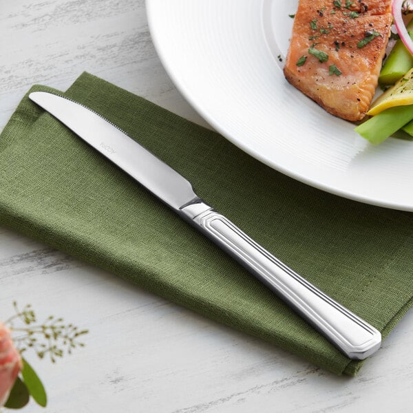 An Acopa Landsdale stainless steel dinner knife on a napkin next to a plate of salmon and vegetables.
