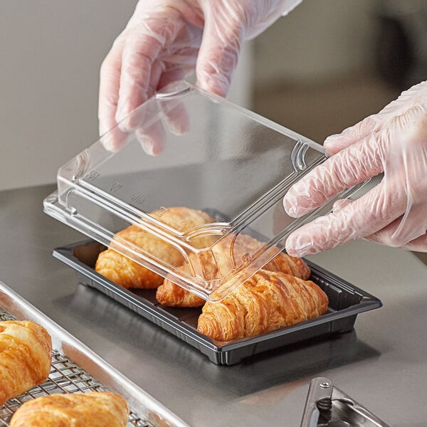 A person in a plastic glove holding a Choice large clear food container with croissants inside.