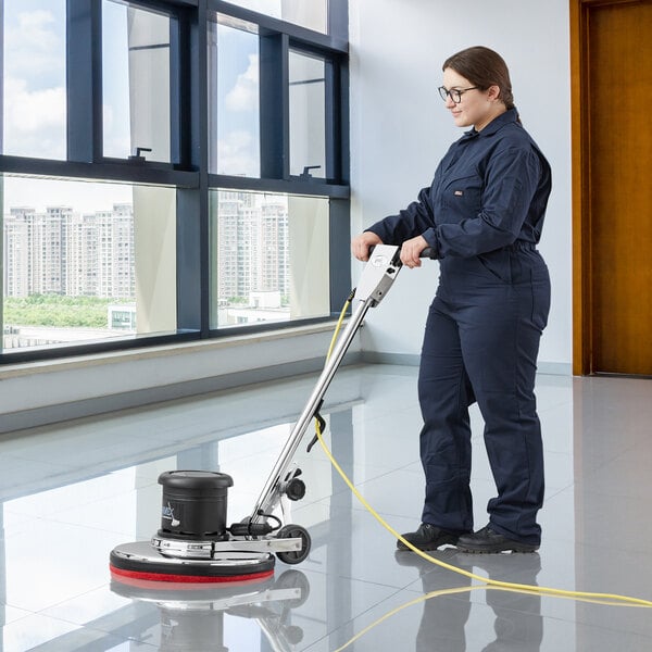 A woman in blue overalls using a Lavex dual speed rotary floor machine to clean the floor in a room.