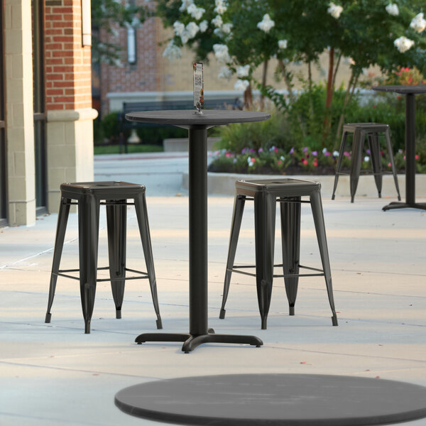 A Lancaster Table & Seating round counter height table with a cross base and three black stools on a patio.