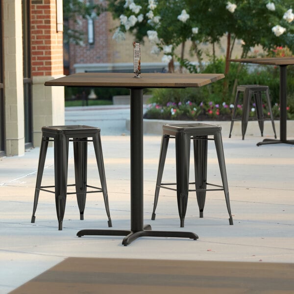 A Lancaster Table & Seating square counter height table with a textured finish and cross base plate on a patio with three black stools.