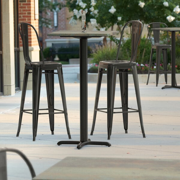 A Lancaster Table & Seating square bar height table with a textured metal finish and cross base plate on a patio with chairs.