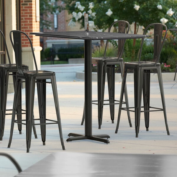 A Lancaster Table & Seating rectangular bar height table with a cross base plate and black chairs on a patio.