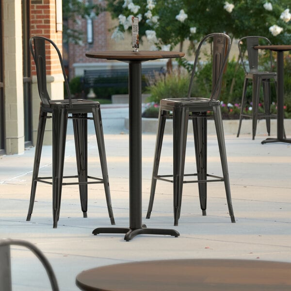 A Lancaster Table & Seating bar height table with a textured brown surface and cross base plate on a patio with black metal bar stools.