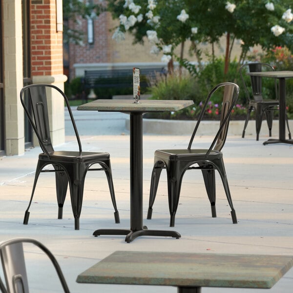A Lancaster Table & Seating square dining table with a textured metal finish on an outdoor patio with black chairs.