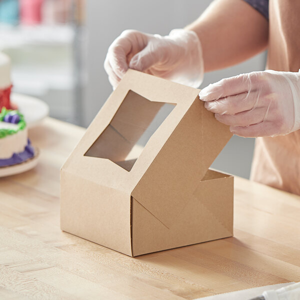 A person in gloves opening a Baker's Mark kraft cardboard box with a window to reveal a cake inside.