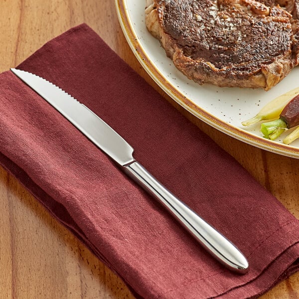 An Acopa Remy stainless steel steak knife on a napkin next to a plate of steak and vegetables.