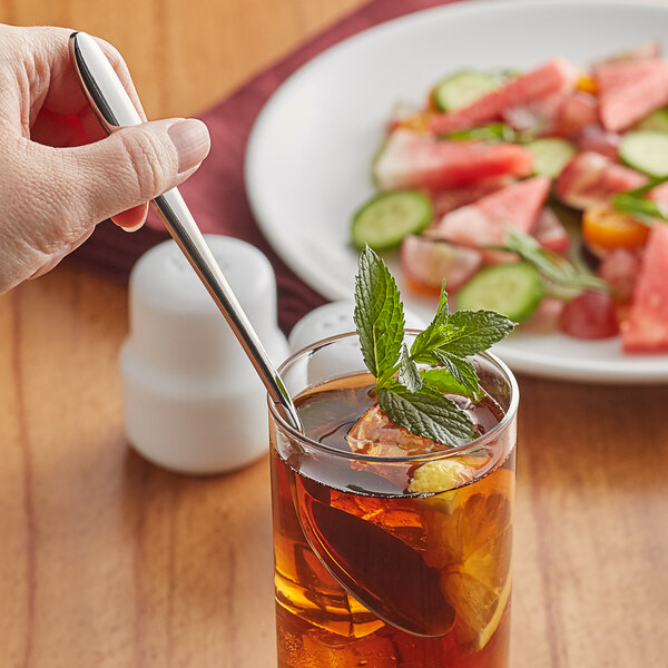 A hand holding an Acopa Remy stainless steel iced tea spoon in a glass of tea with mint leaves.