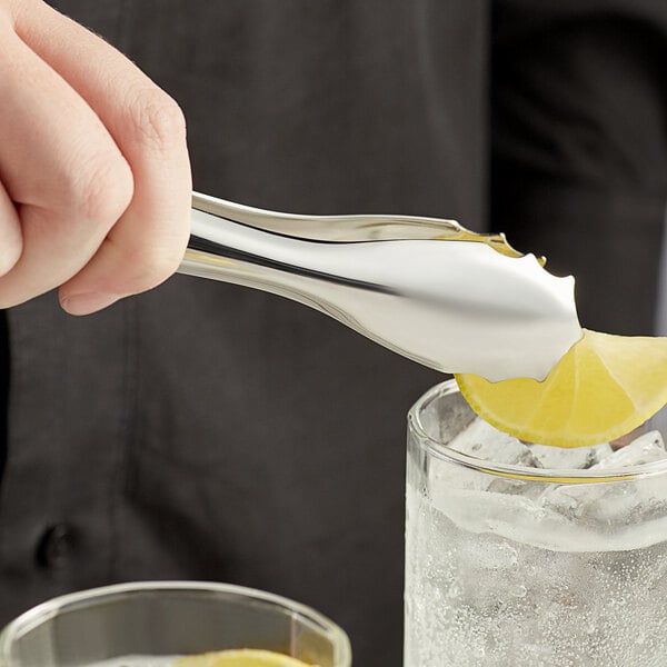 A person using Acopa stainless steel tongs to squeeze a lemon wedge into a glass of ice.