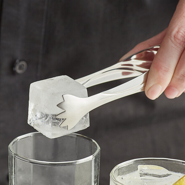 A person using Acopa stainless steel tongs to pick up an ice cube in a glass.