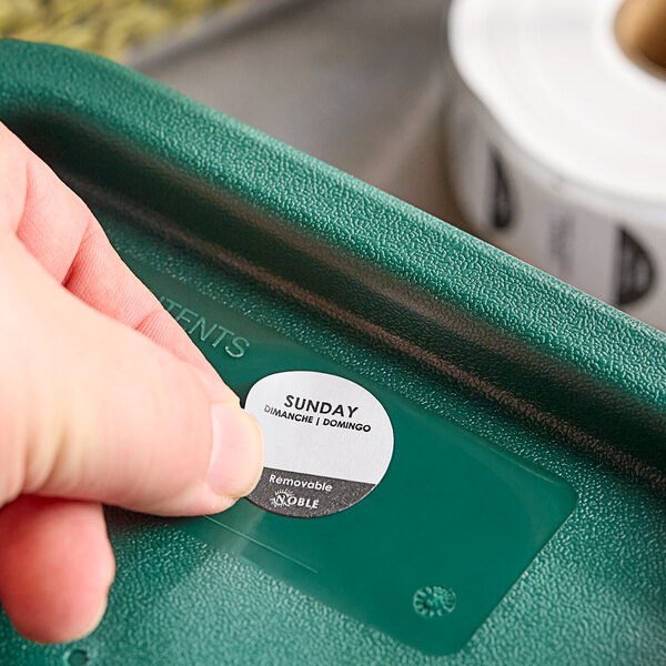 A hand putting a Noble Products Sunday label on a green plastic container.