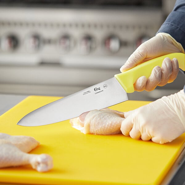 A person in gloves using a Choice chef knife with a neon yellow handle to cut chicken on a yellow cutting board.