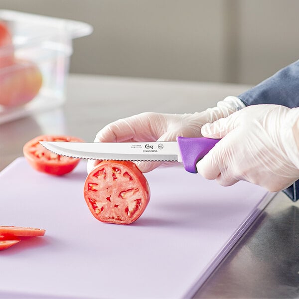 A person in gloves using a Choice serrated utility knife with a purple handle to cut a tomato.