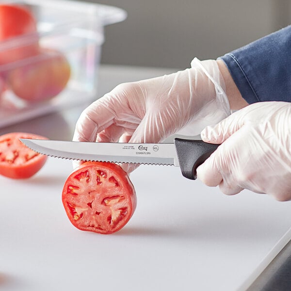 A person using a Choice serrated utility knife with a black handle to cut a tomato.