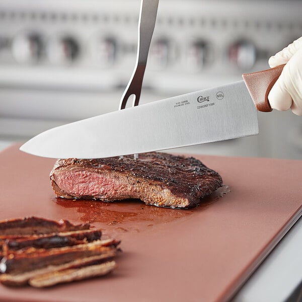 A person using a Choice 10" chef knife with a brown handle to cut a steak on a cutting board.