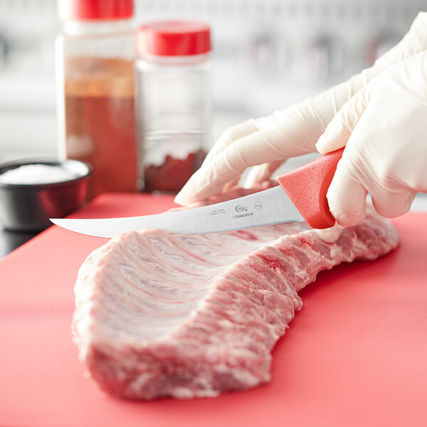 A person using a Choice curved stiff boning knife with a red handle to cut a piece of meat on a red cutting board.