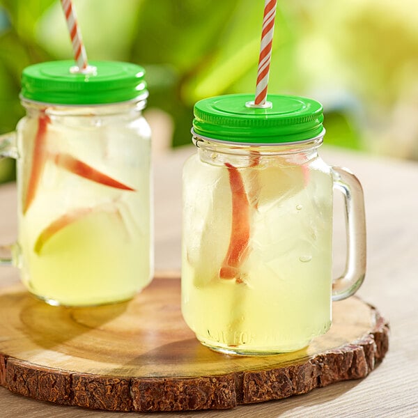 Two Acopa glass jars with green metal lids and straws filled with a yellow drink.
