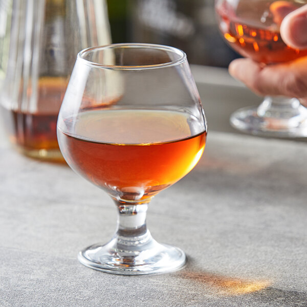 A hand holding a Pasabahce Capri brandy glass filled with brown liquid.