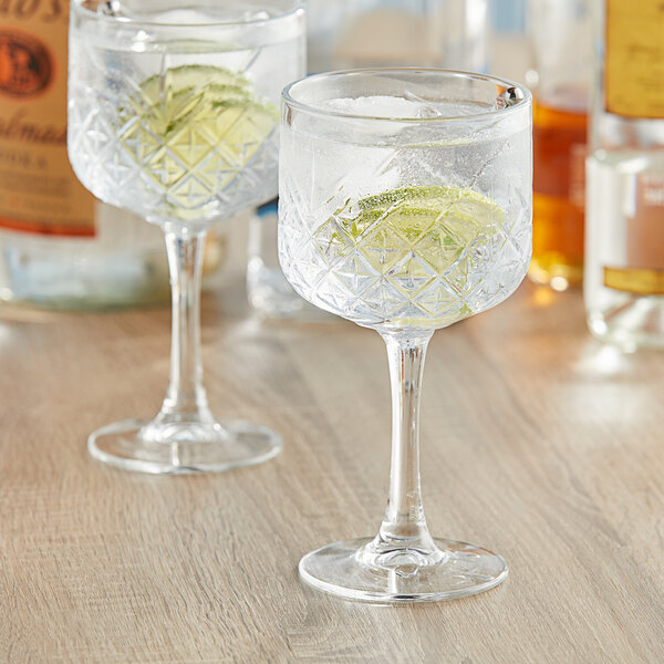 Two Pasabahce Timeless Vintage gin and tonic glasses filled with ice and lime slices on a wooden table.