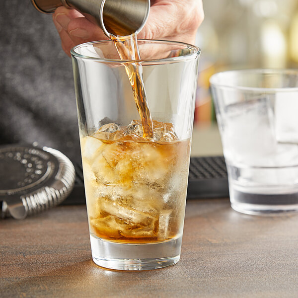 A person pouring brown liquid into a Pasabahce mixing glass of ice cubes.