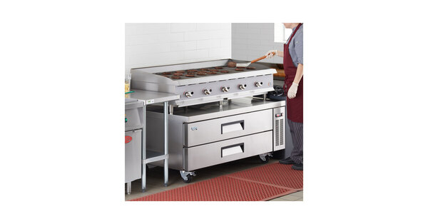 A woman in a red apron cooking food on a large gas grill over a chef base.