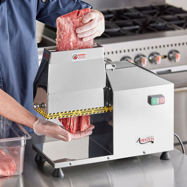 A person in a blue uniform using an Avantco meat tenderizer to cut meat on a counter.