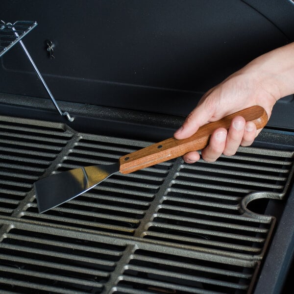 A person using an American Metalcraft stainless steel slanted edge grill scraper with a long wooden handle to clean a grill.