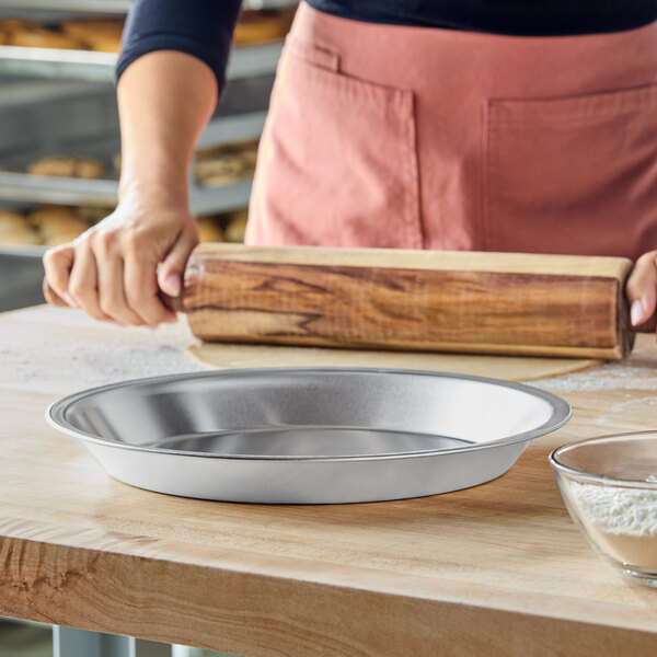 A person is rolling out dough with a wooden rolling pin on a floured surface next to an empty metal pie pan and a glass bowl of flour.