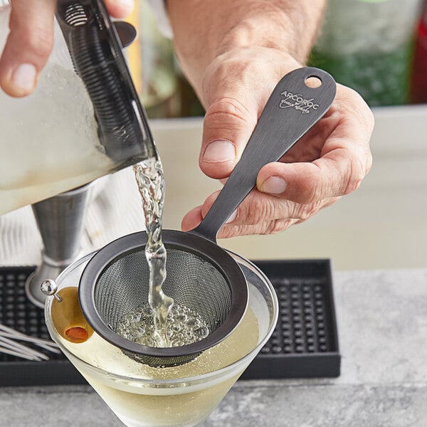 A person using an Arcoroc matte black stainless steel fine mesh strainer to pour a martini.