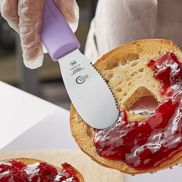 A Mercer Culinary purple scalloped sandwich spreader being used to spread jam on a piece of bread.
