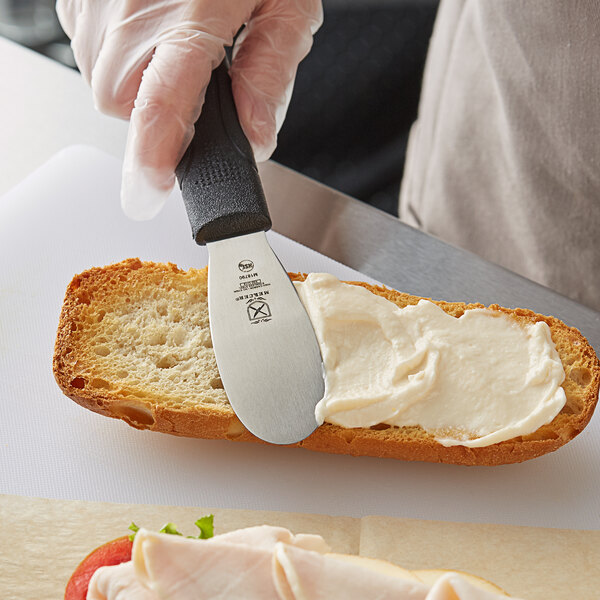 A person's hand with a Mercer Culinary black handle knife spreading cream on a piece of bread.