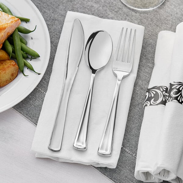A white napkin with a Hoffmaster metallic silver plastic fork and knife on top of it on a table with a plate of food.
