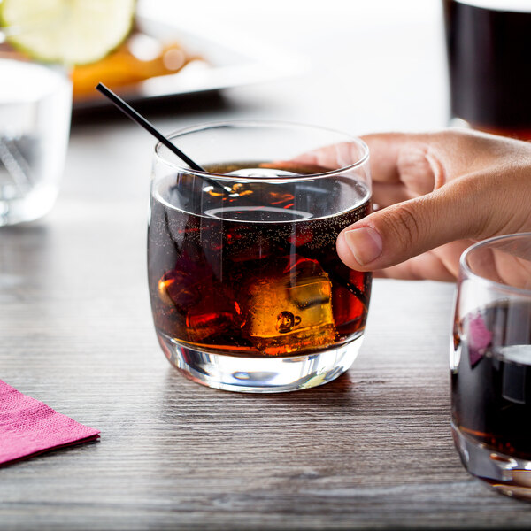 A hand holding a Chef & Sommelier Cabernet rocks glass filled with brown liquid and ice with a straw.