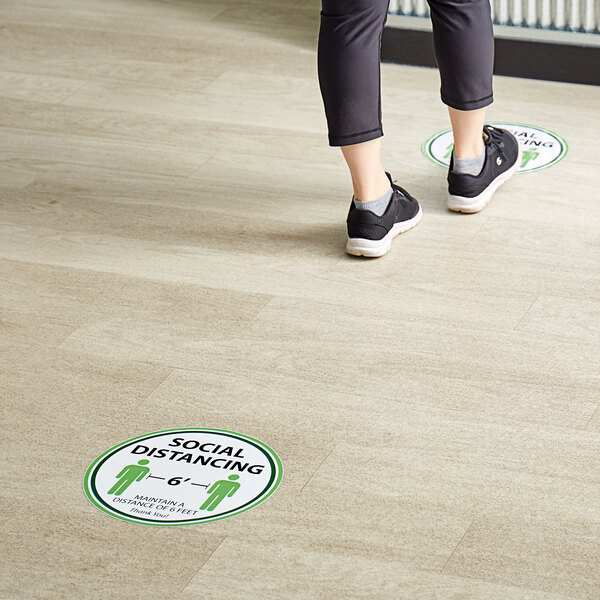 A person standing on a floor with a Tablecraft "Social Distancing" floor decal.